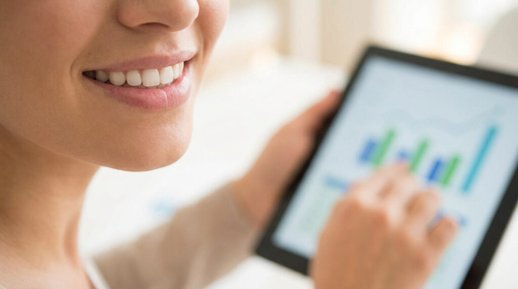 Gros plan sur une femme souriante, les dents blanches. Elle regarde une tablette affichant des graphiques flous qu'elle touche du doigt.